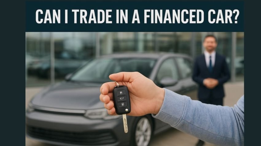 A person holding a car key fob in front of a gray sedan at a dealership, symbolizing trading in a financed car. The text overlay reads “Can I Trade In a Financed Car?”, representing the process of trading a car that’s still under loan.